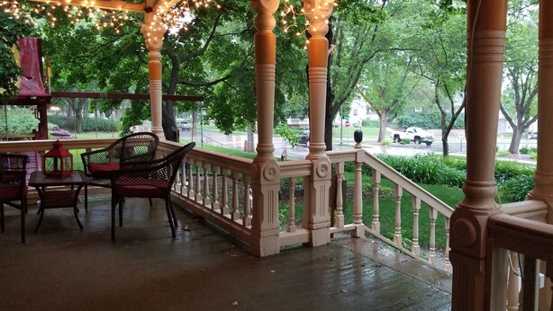 A covered outdoor area with a wooden railing, chairs, and tables. - Historic Victorian Inn (Sioux Falls)