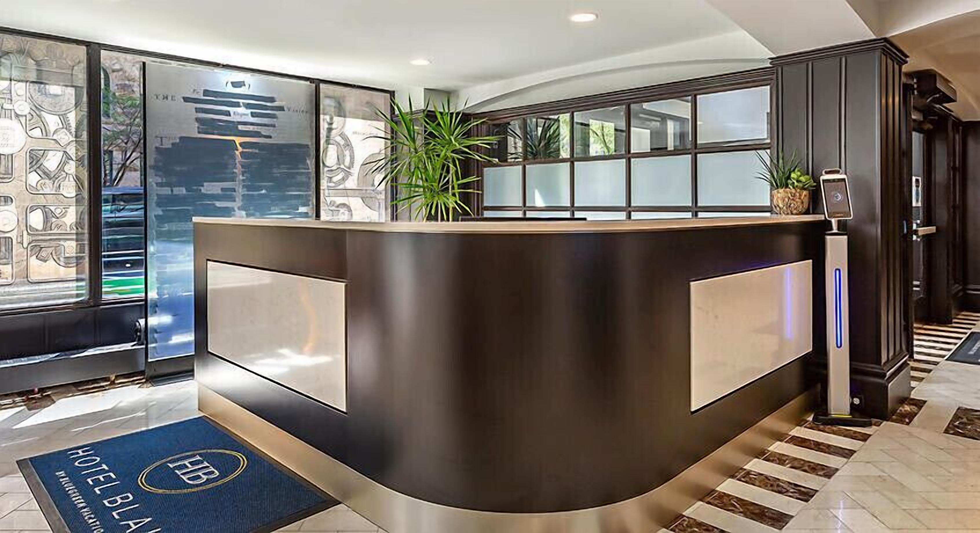 A modern hotel reception desk with a dark wood finish, glass panels, and a blue mat with the hotel logo.