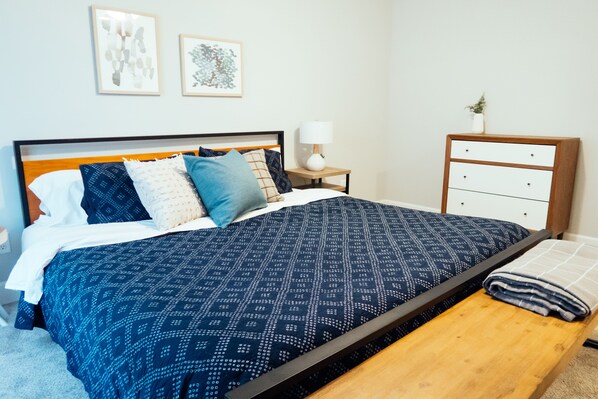 A neatly made bed with a patterned blue and white duvet, a wooden bench, a bedside table with a lamp, and a dresser with a plant.