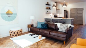 A modern living room with a brown leather sofa, a white coffee table, and a yellow armchair. There is a kitchen area with white cabinets and stainless steel appliances.