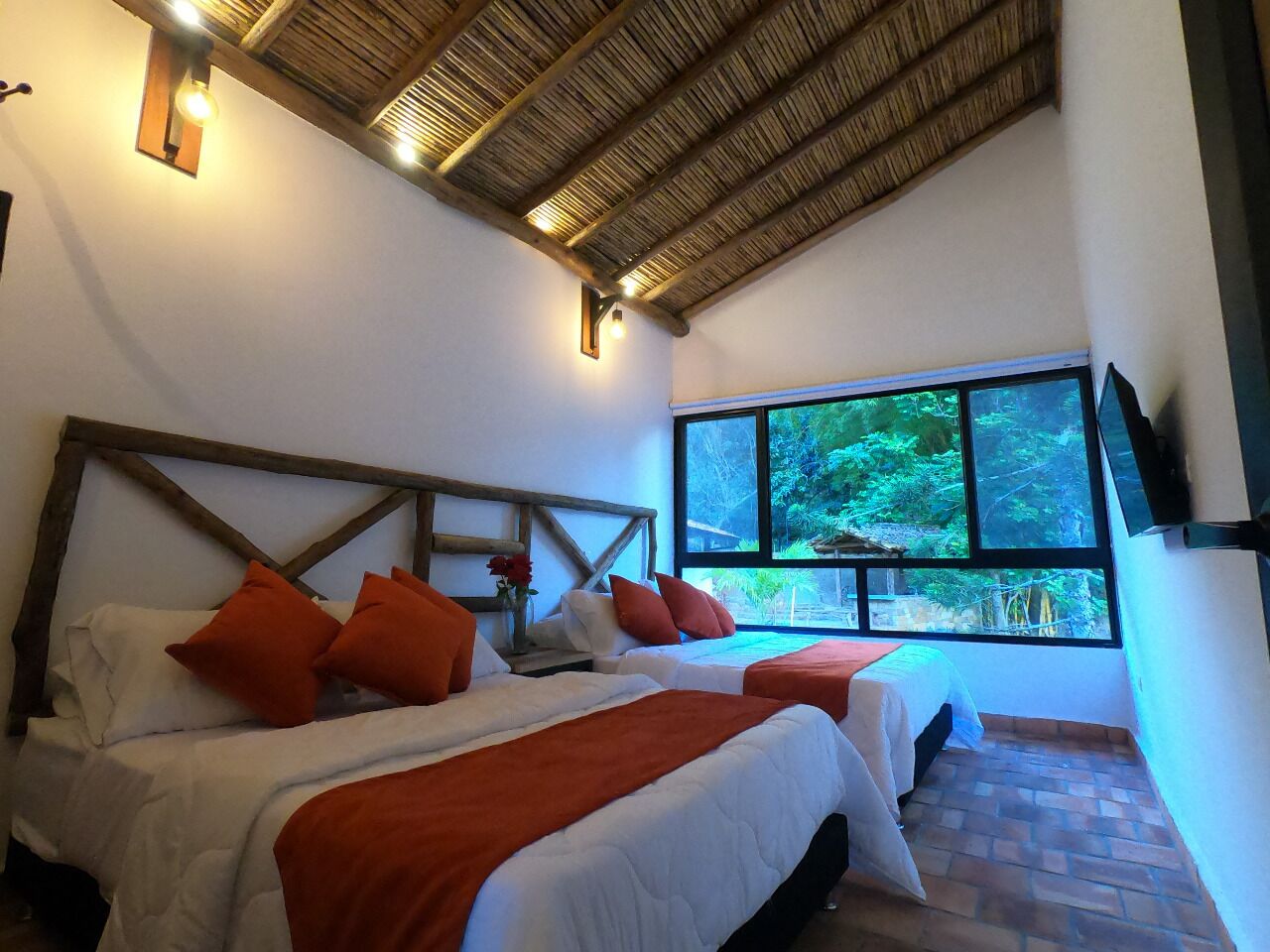 A bedroom with two beds, a large window with a view of greenery, a wooden ceiling, and a wall-mounted TV.