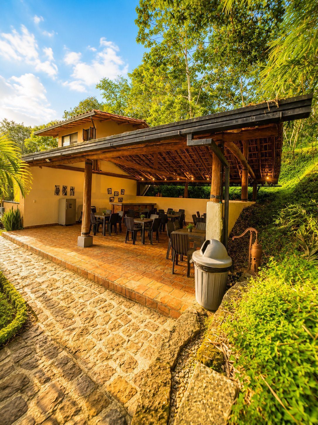 A covered outdoor seating area with tables and chairs, a trash bin, and a stone pathway leading to a building.
