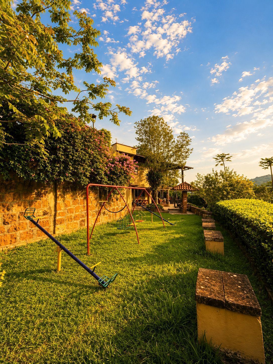 A grassy outdoor area with a swing set, a small brick structure, and a stone wall.