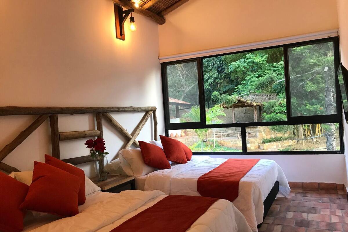 Ein Zimmer mit zwei Betten, roter und weißer Bettwäsche, einem Holz-Kopfteil, einem großen Fenster mit Blick auf Grünflächen und einem Ziegelboden.