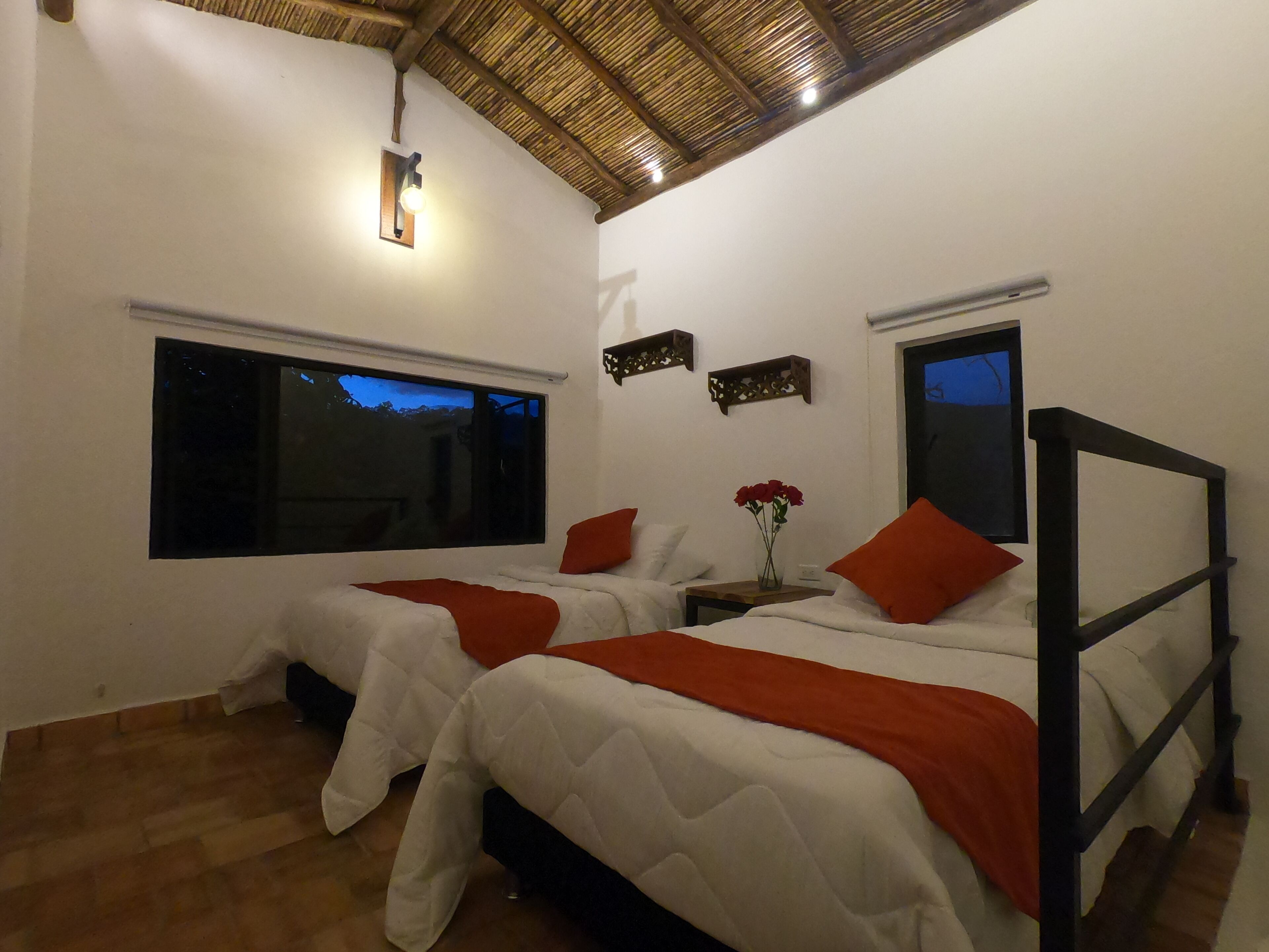 A bedroom with two beds, a large window, a small table with a vase of flowers, and a wooden ceiling.