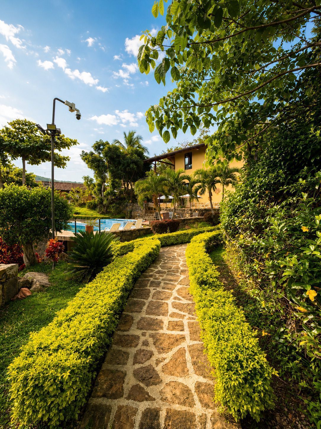 A stone pathway leading to a pool area, surrounded by lush greenery and a yellow building.