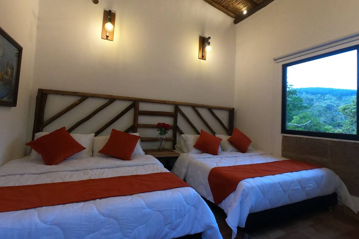 Ein Hotelzimmer mit zwei Betten, jeweils mit weißer Bettwäsche und roten Kissen, einem Holzkopfstück und einem großen Fenster mit Blick auf eine bewaldete Landschaft.