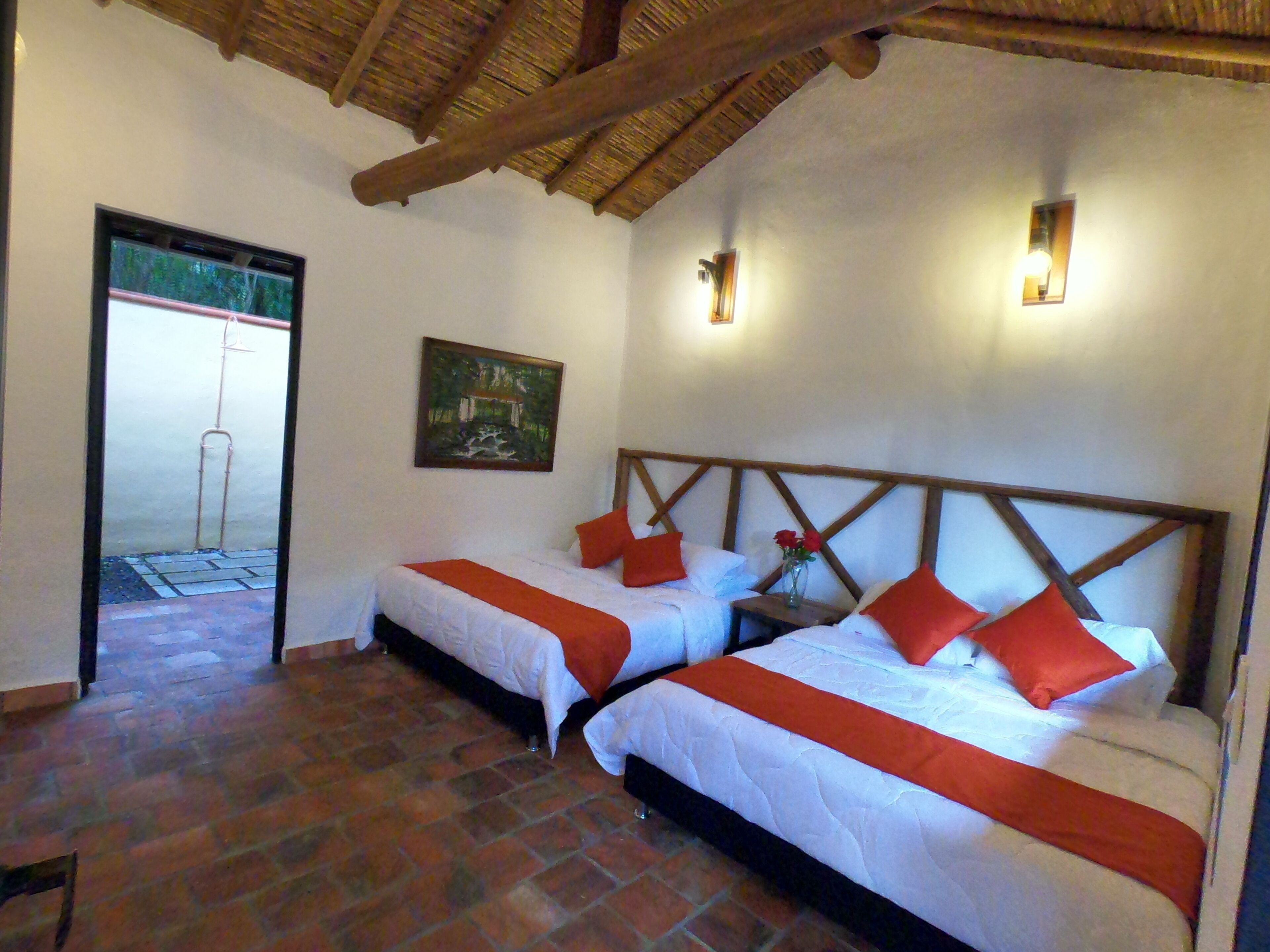 A room with two beds, each with white bedding and orange accents, a wooden headboard, a painting on the wall, and a small table with a vase of flowers.