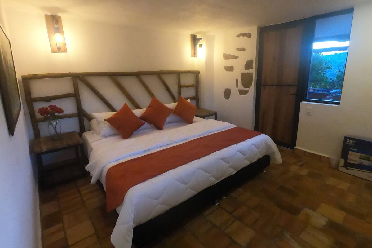 Ein Schlafzimmer mit einem hölzernen Kopfteil, einem Bett mit weißer Bettwäsche und orangenen Kissen, einem Nachttisch mit Blumenvasen und einem Fenster mit Blick ins Grüne.