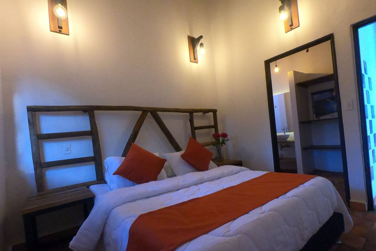 Ein ordentlich bezogenes Bett mit weißer und orangefarbener Tagesdecke, neben zwei Nachttischen mit Lampen, in einem Zimmer mit Holz-Kopfschild und Spiegel an der Wand.