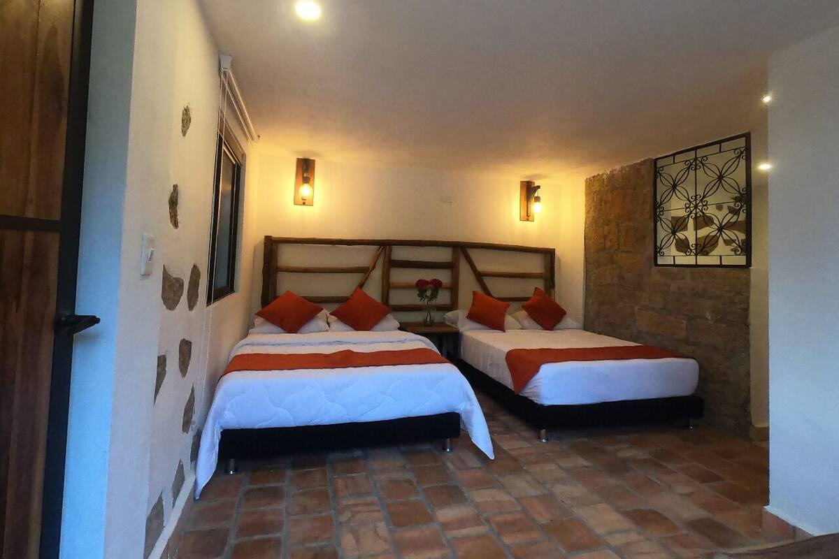 Ein Zimmer mit zwei Betten, jeweils mit weißer Bettwäsche und orangefarbenen Kissen, einer Steinwand und einem dekorativen Metallkunstwerk an der Wand.