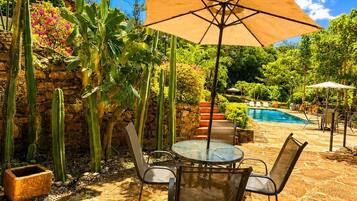 Un patio con piscina, rodeado de cactus y palmeras, con una mesa y sillas bajo un gran paraguas.