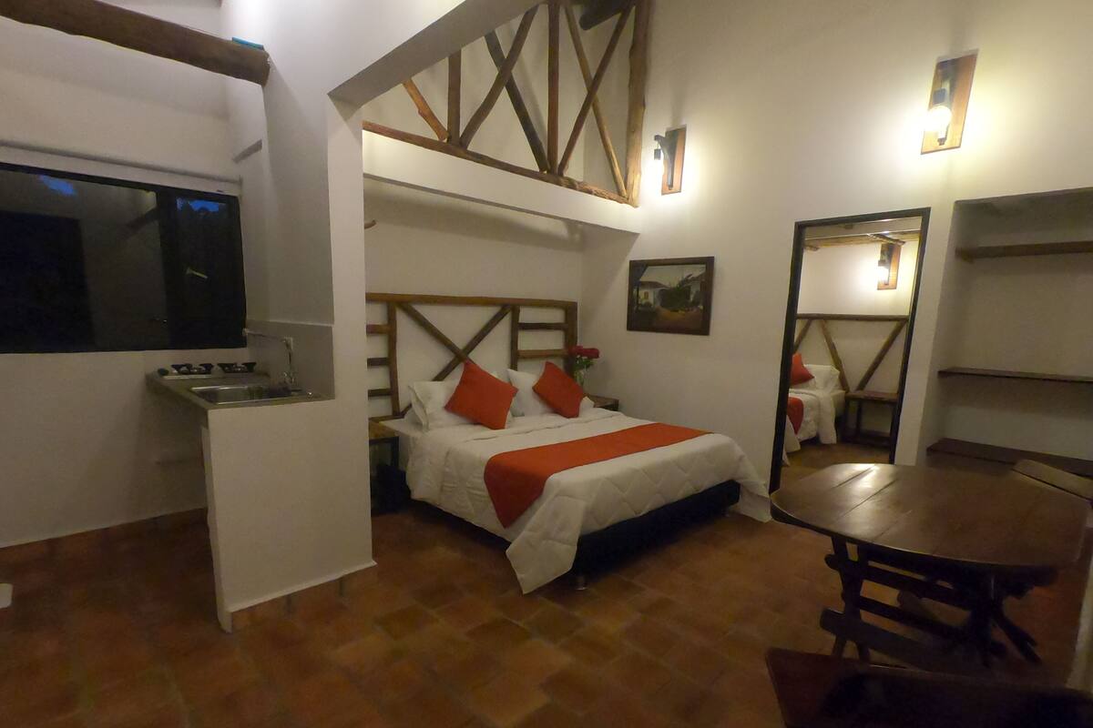 Ein kleines Hotelzimmer mit Bett, einer kompakten Küchenzeile, einem Esstisch und einem Spiegel.