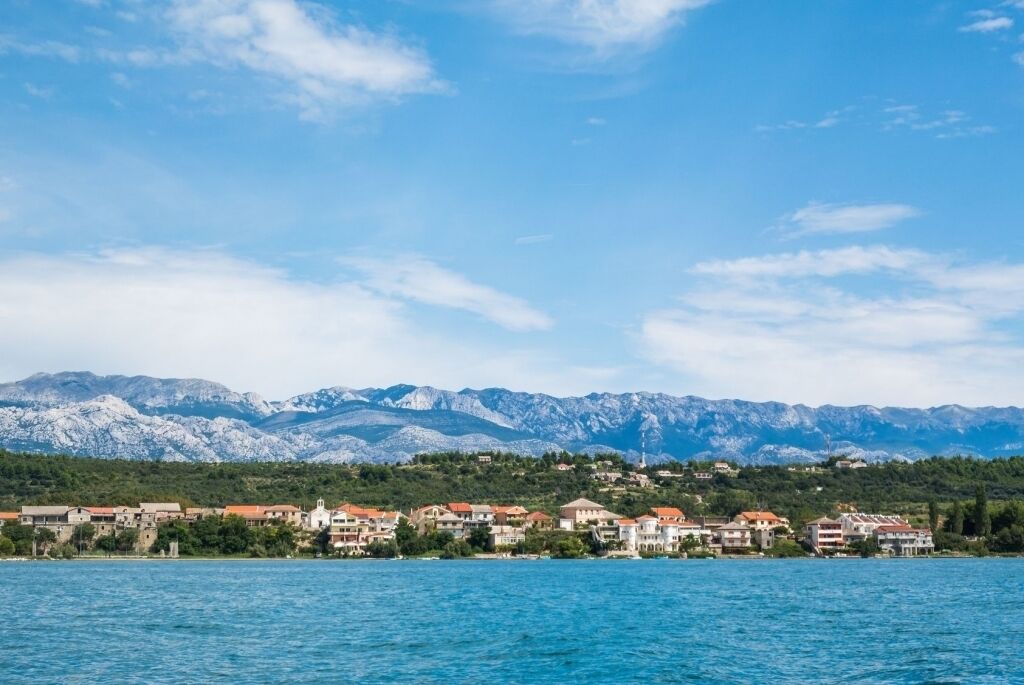 A coastal town with buildings along the shoreline, a clear blue sky, and mountains in the background.