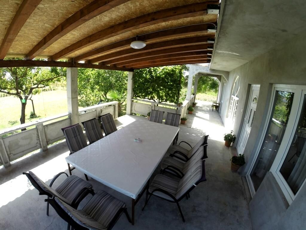 An outdoor patio with a large table and chairs, a covered area with a wooden ceiling, and a view of the surrounding greenery.