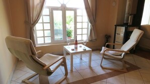 A room with two lounge chairs, a small table with a bottle and glasses, and a view through a window with curtains.