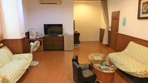 Une chambre d’hôtel avec deux canapés à motifs, une petite table avec un plateau en verre, un téléviseur, un mini-réfrigérateur, un ventilateur et une armoire en bois.