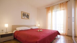 A neatly made bed with a red bedspread, two bedside tables with lamps, a framed picture on the wall, and a window with curtains.