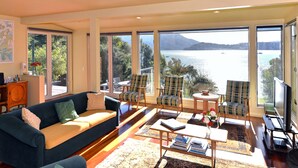 A spacious living room with a large sofa, two armchairs, a coffee table, and a television. The room has large windows offering a view of a lake and mountains. - Ngaire's Haven - Mahau Sound Holiday Home (Mahau Sound)
