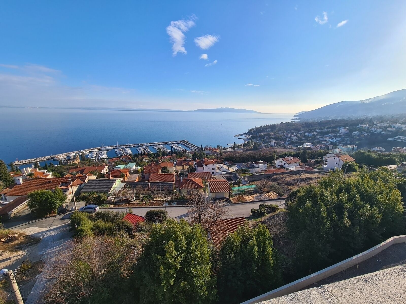 A coastal town with a marina, houses with red-tiled roofs, and a view of the sea and distant mountains.