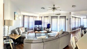 Amplia sala de estar con un sofá grande, una mesa de centro y un televisor. Hay un comedor con mesa y sillas.