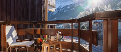 A balcony with a wooden chair and a small table, overlooking a snowy mountain landscape.