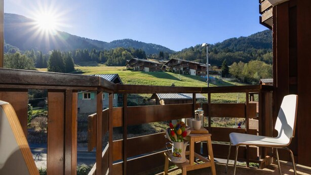 Un balcón con barandilla de madera, una silla blanca, una mesita con un jarrón de flores y vistas a casas y montañas al fondo.