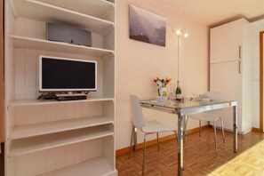 Een moderne woonkamer met een flatscreen televisie op een witte plank, een glazen eettafel met stoelen, en een schilderij aan de muur.