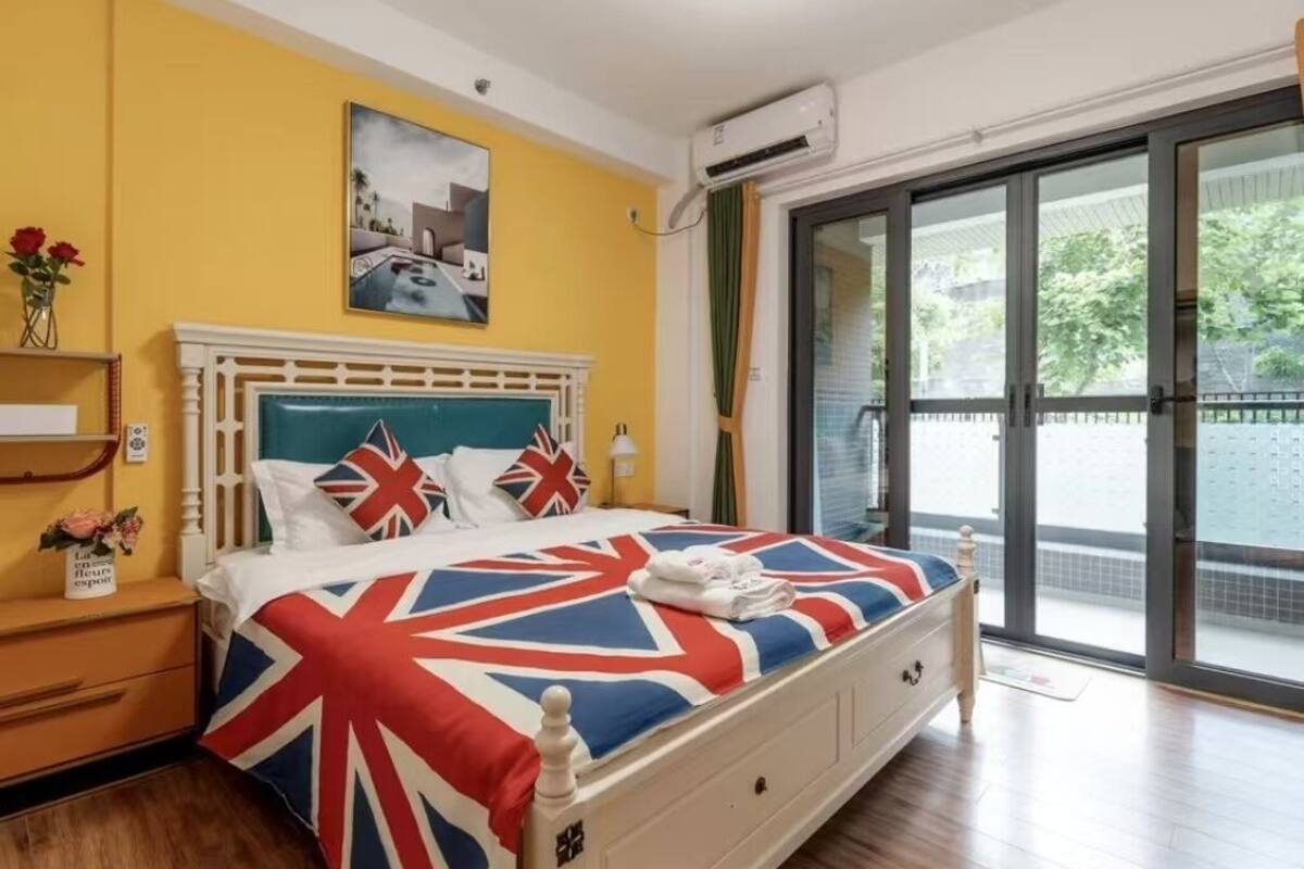 A bedroom with a bed featuring a Union Jack bedspread, a white headboard, and a nightstand with a vase of flowers.