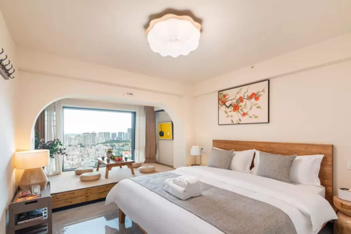 A hotel room with a large bed, a desk with a lamp, a view of the city through a window, and a ceiling light.