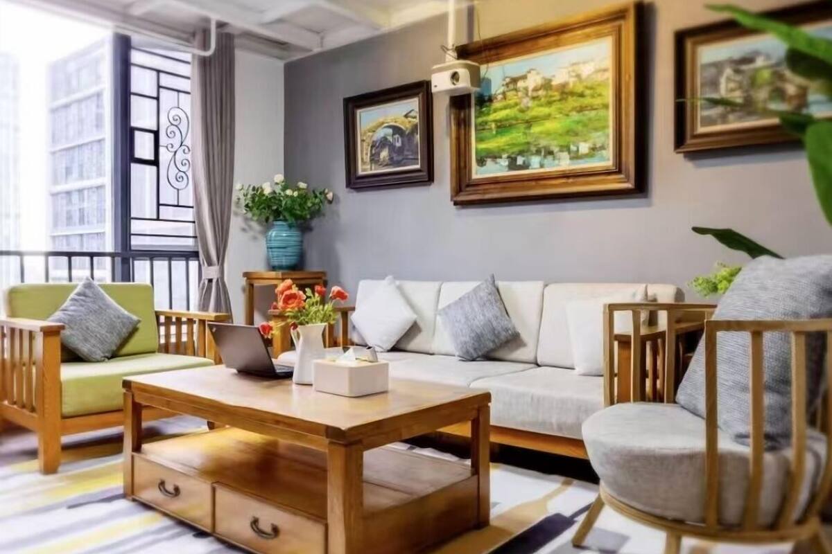 A modern living room with a sofa, two armchairs, a wooden coffee table, and a laptop. There are framed pictures on the wall, a potted plant, and a window with a view of buildings.