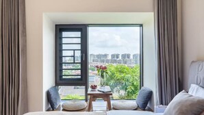 Ein Schlafzimmer mit großem Fenster, einem Bett, einem kleinen Tisch mit Blumenvase und Blick auf eine Stadtlandschaft.