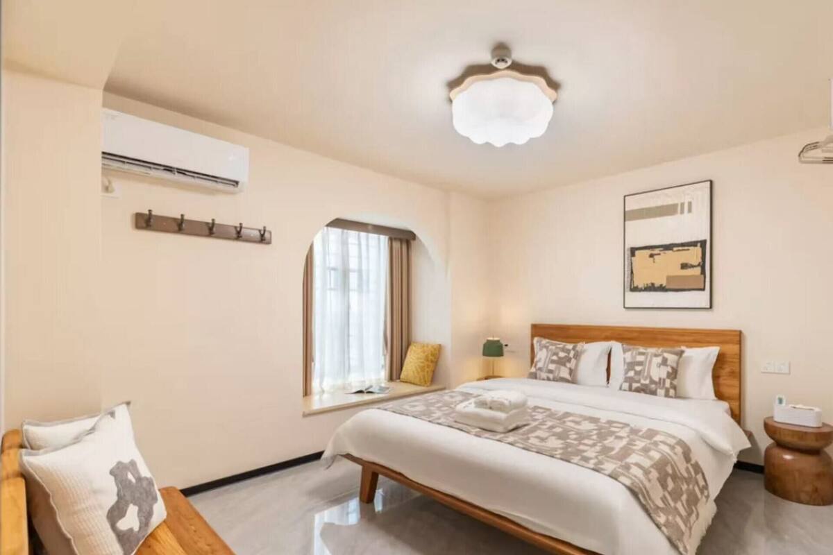 A hotel room with a bed, a window, a wall-mounted air conditioner, a bedside lamp, and a small wooden stool.