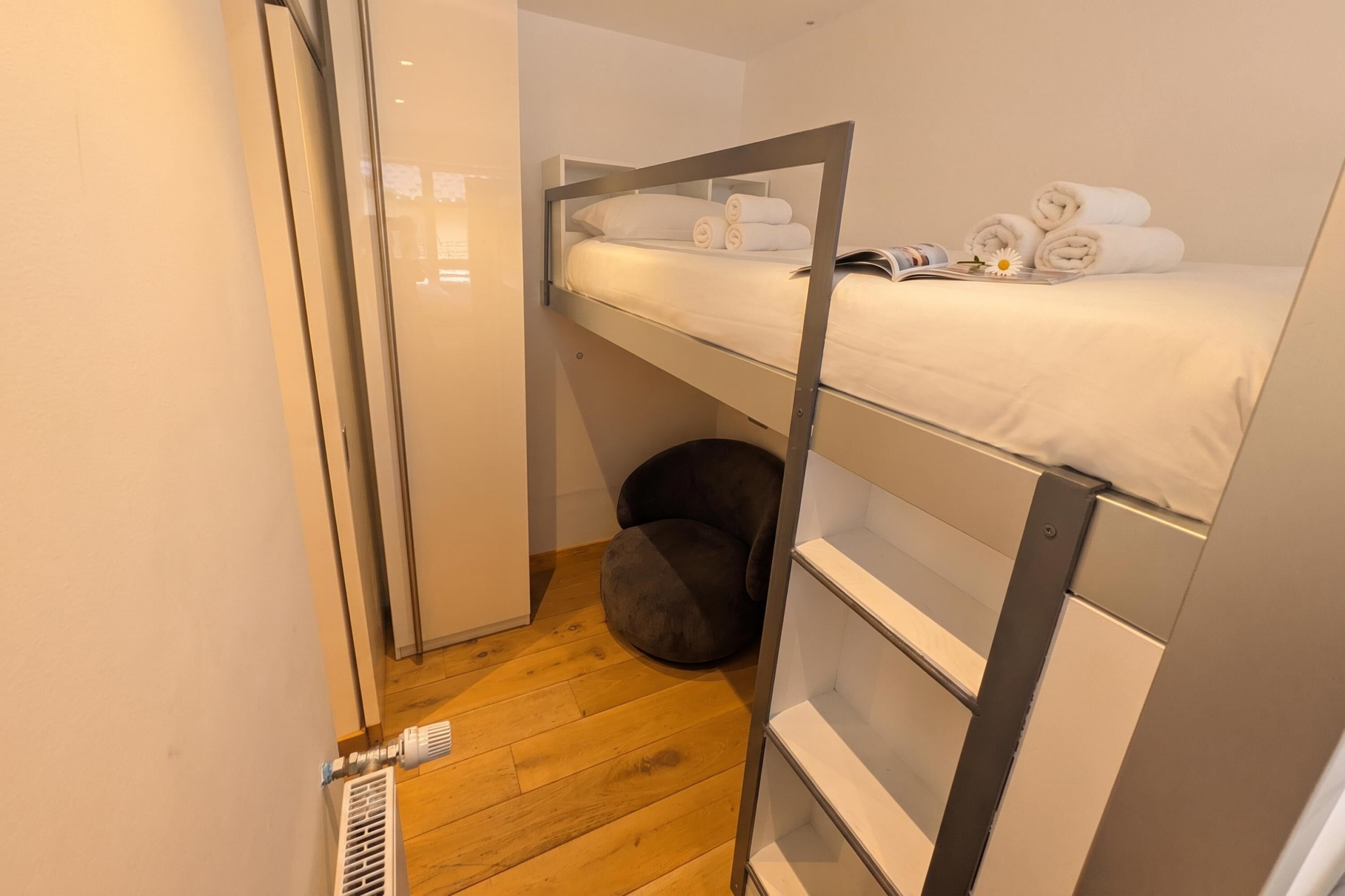 A bunk bed with a lower shelf, a small round ottoman, and a radiator.