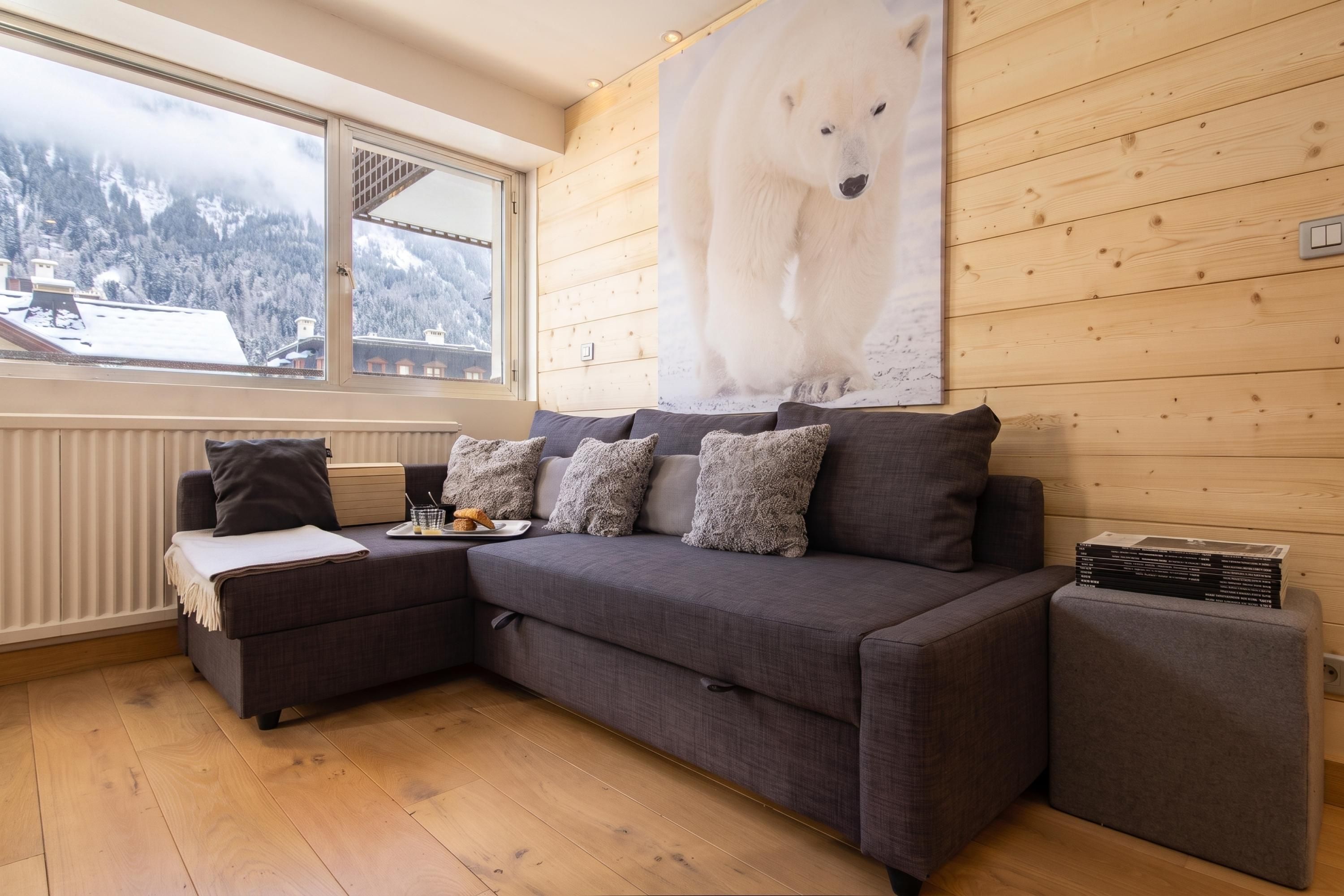 Ein gemütliches Zimmer mit einer grauen Couch, einem kleinen Beistelltisch mit einer Croissantplatte und einem Fenster mit Blick auf einen verschneiten Berg.