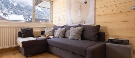A cozy room with a grey sofa, a small side table with a tray of croissants, and a window overlooking a snowy mountain.