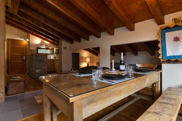 Una mesa de comedor de madera con cubierta de vidrio, rodeada de sillas, en un interior rústico con vigas de madera a la vista y una chimenea de piedra.