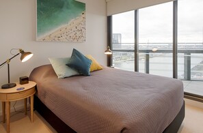 A bedroom with a large bed, a nightstand with a lamp, a painting of a beach, and a view of a cityscape through large windows. - Orange Stay Premier (Docklands)