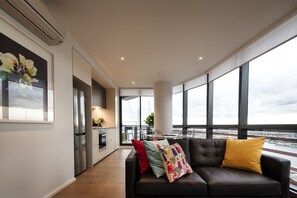A modern living room with a black leather sofa, colorful cushions, and large windows offering a view of a cityscape. - Orange Stay Premier (Docklands)