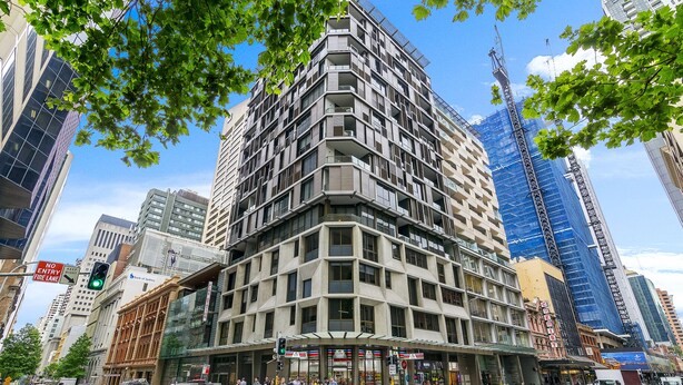 Un immeuble moderne de grande hauteur, avec une entrée située à l’angle, entouré d’autres bâtiments imposants et bordé par une rue animée.