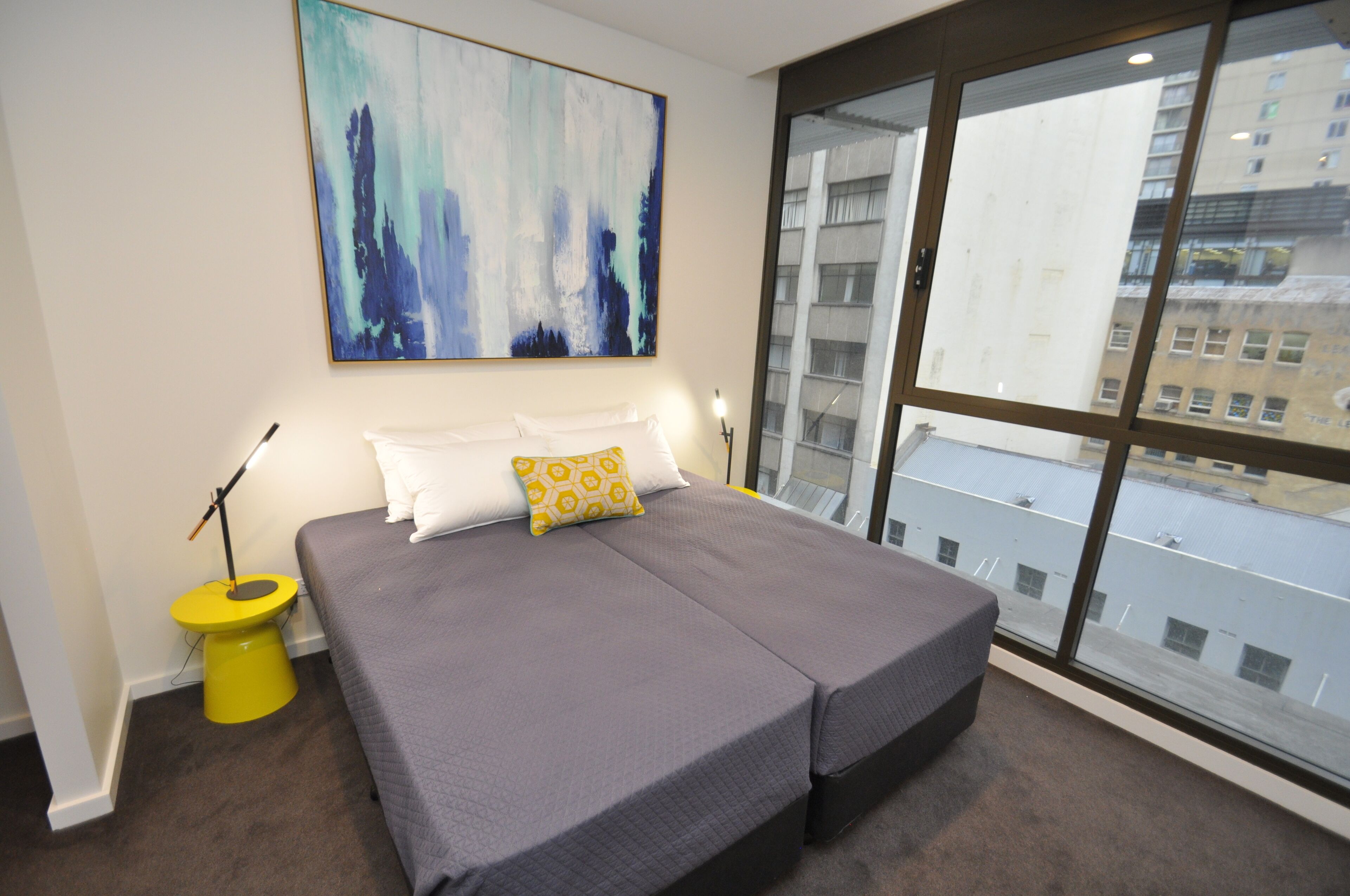 Ein Schlafzimmer mit einem großen Bett, einem gelben Nachttisch, einem Bild an der Wand und Blick auf Gebäude durch das Fenster.