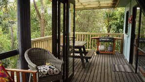 Una terraza de madera con una silla de mimbre y una mesa, una parrilla y vistas de exuberante vegetación.