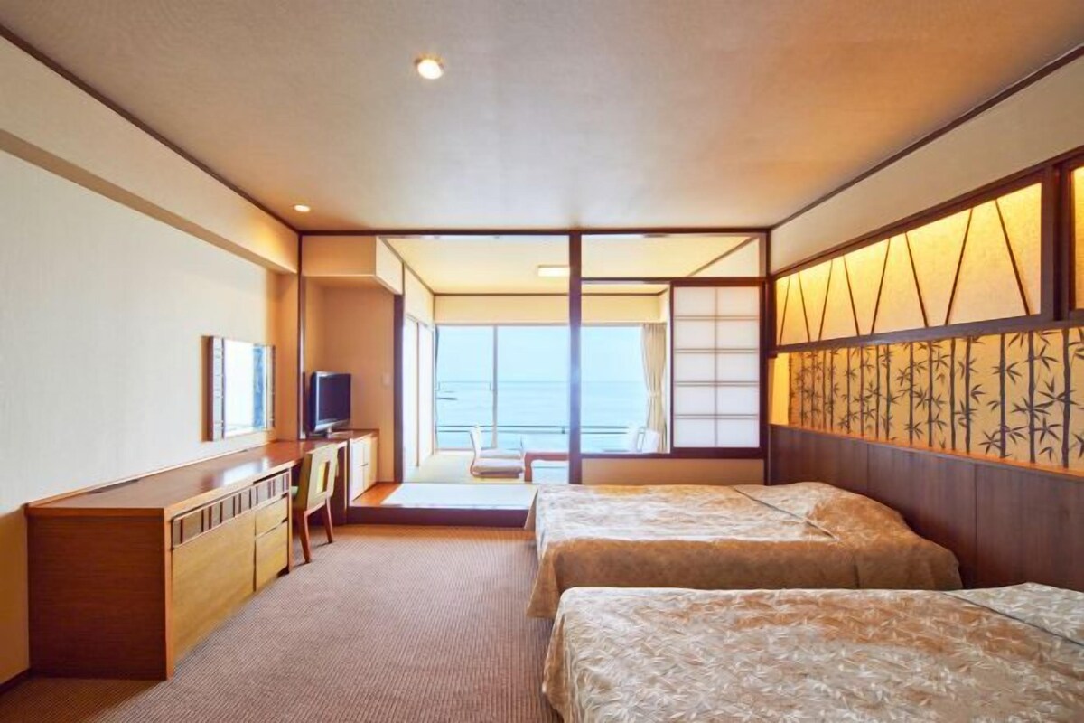 A hotel room with two beds, a desk, a television, and a view of the sea.