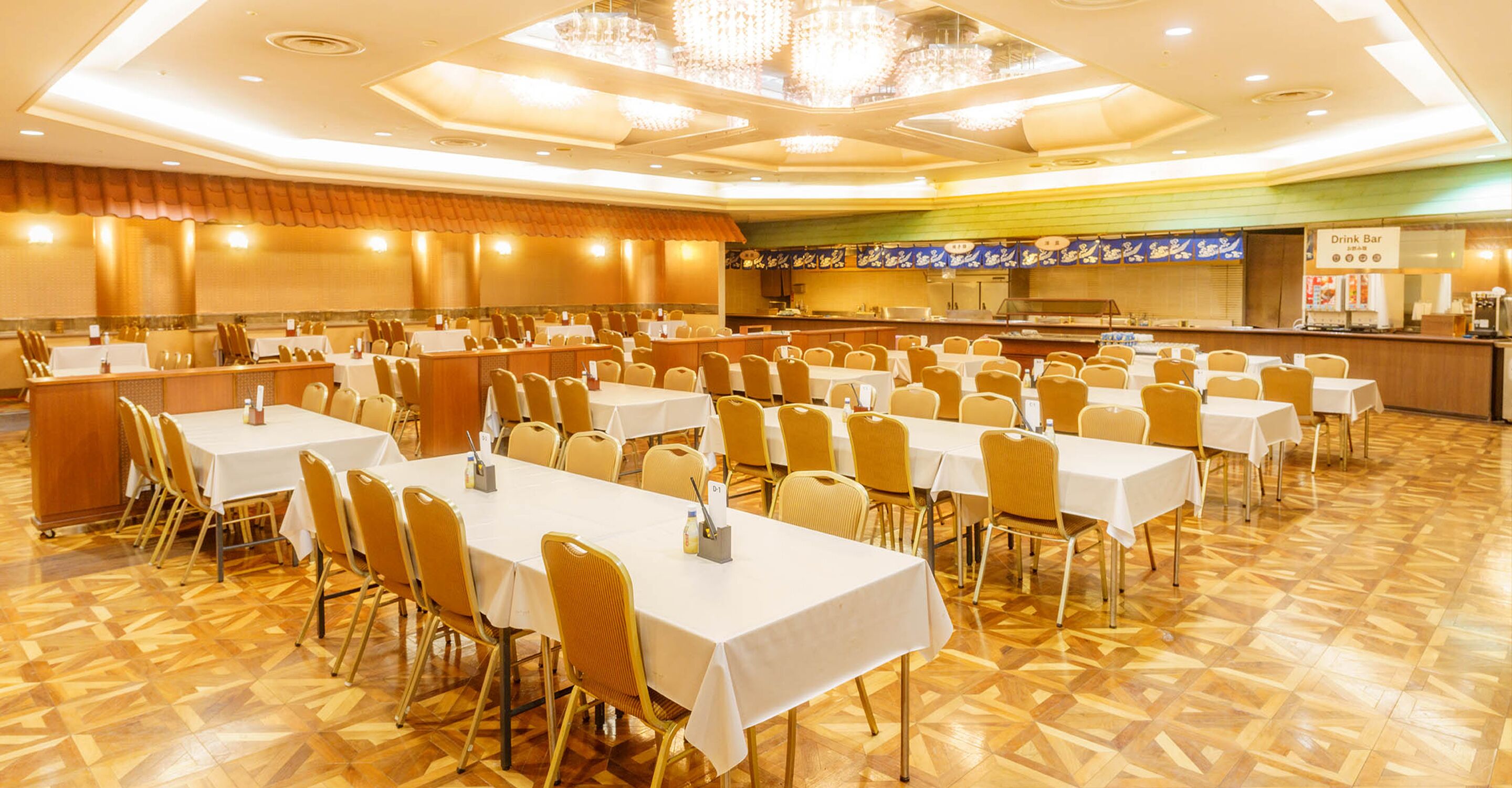 A banquet hall with multiple tables and chairs, a kitchen area, and a drink bar.