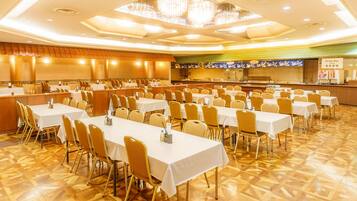 A banquet hall with multiple tables and chairs, a kitchen area, and a drink bar.