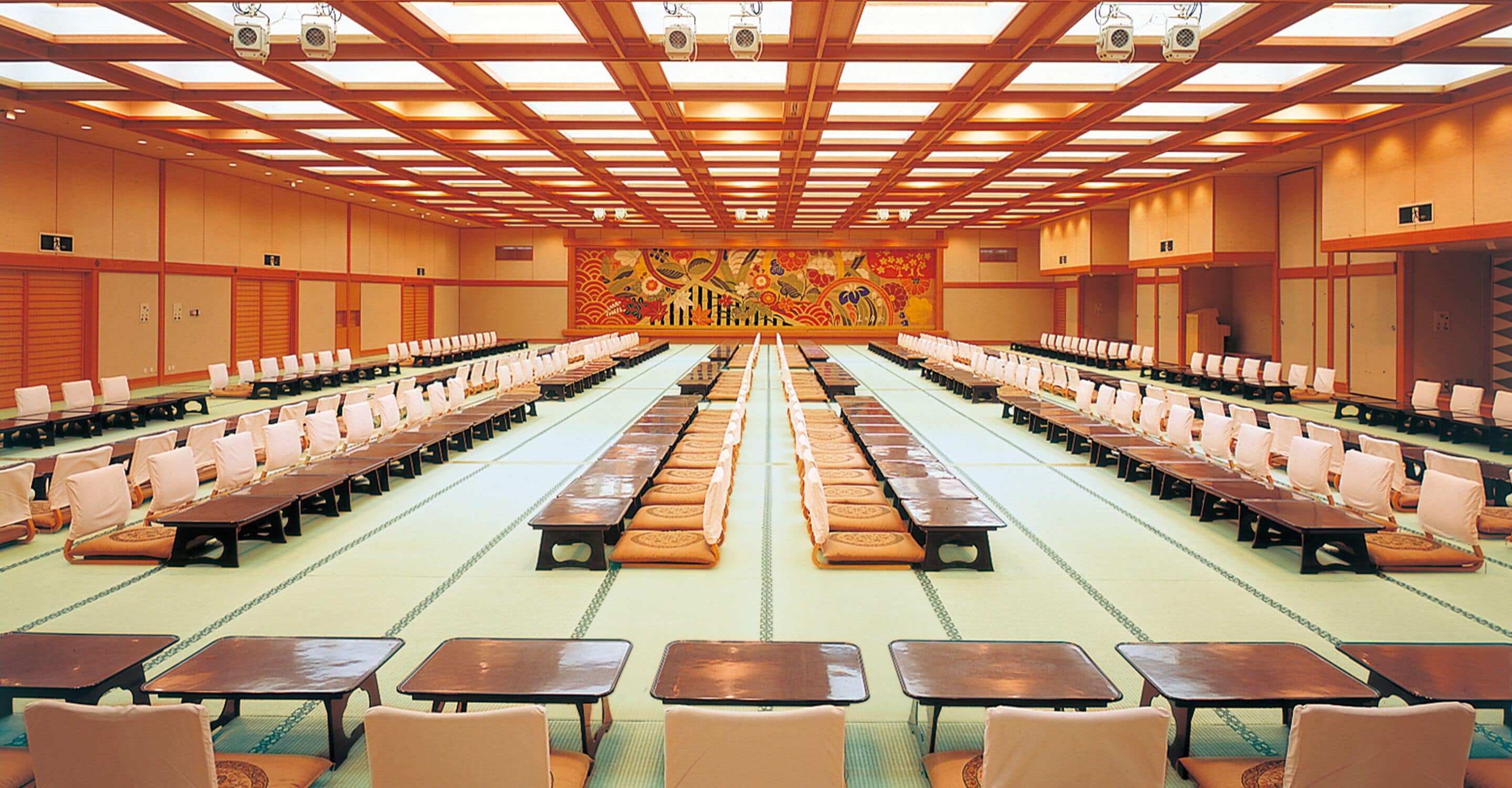 Un amplio salón de conferencias con filas de mesas y sillas, un techo alto con vigas de madera y un mural colorido en la pared de fondo.