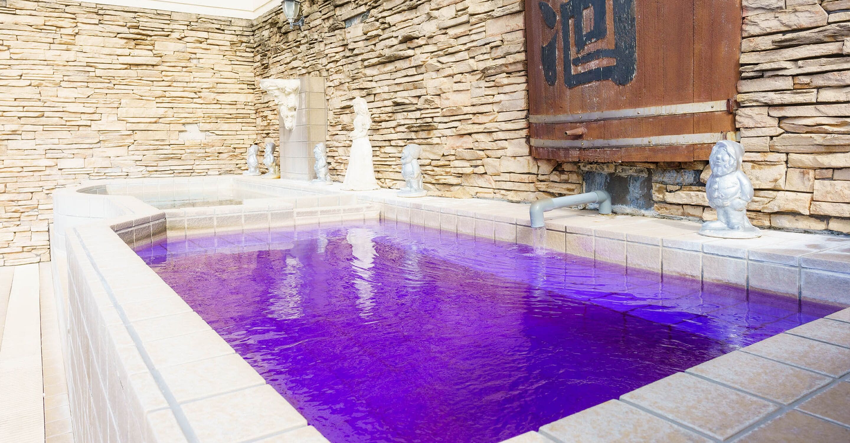 A stone-walled pool with a purple hue, surrounded by stone tiles and featuring a wooden sign with Chinese characters.