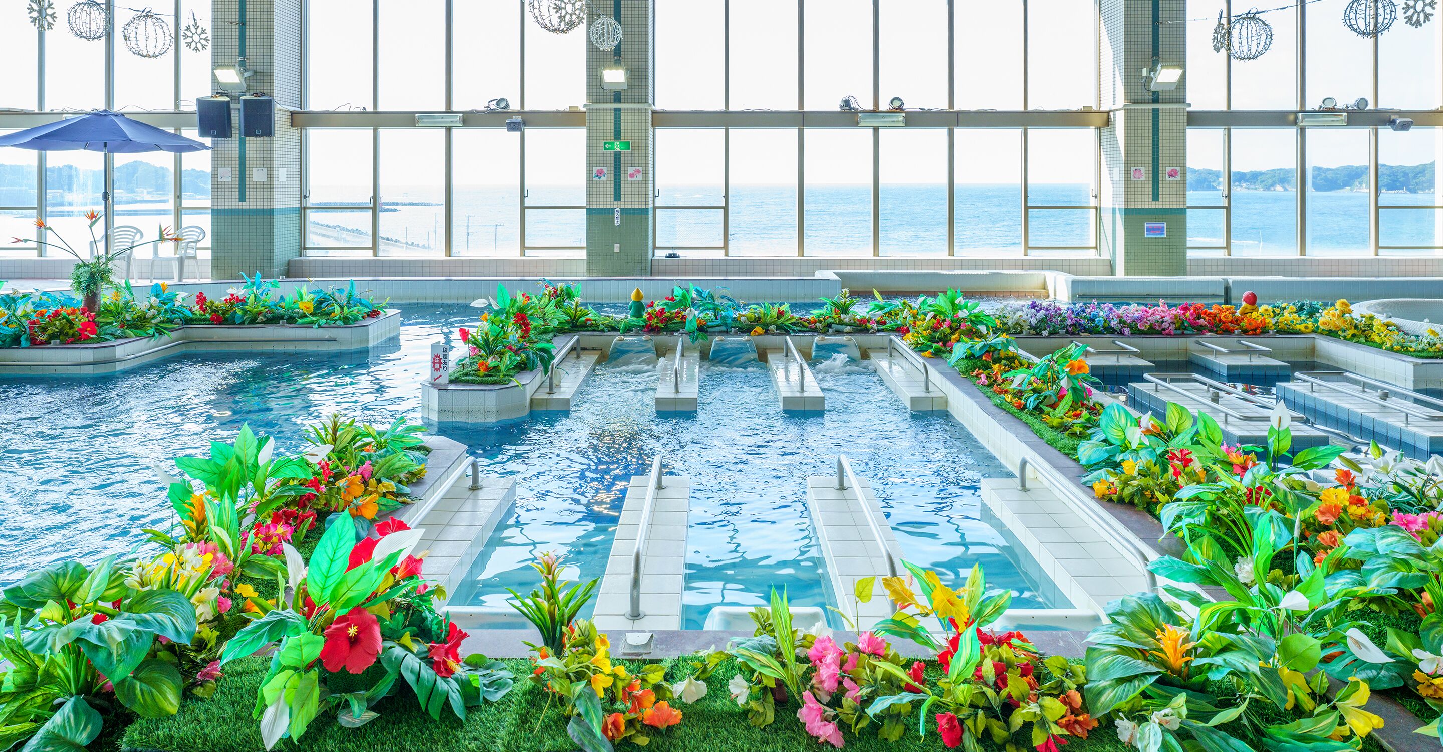 En simbassäng med flera banor, omgiven av färgstarka blomsterrabatter och stora fönster som erbjuder utsikt över havet.