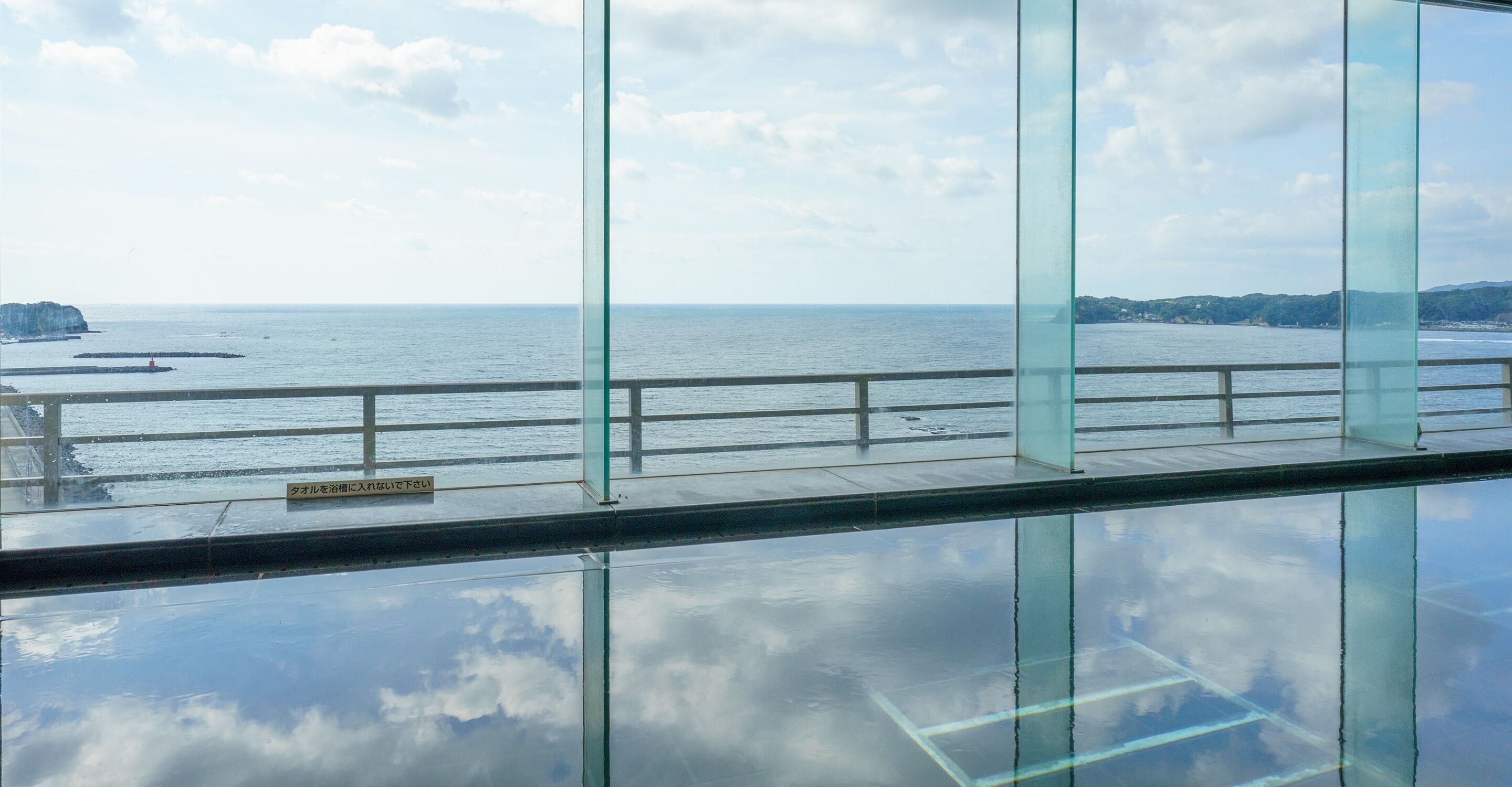 Vista da interno di un edificio con ampie vetrate che si affaccia su una zona costiera, con cielo sereno e mare calmo.