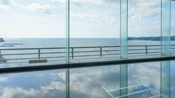 Vista da interno di un edificio con ampie vetrate che si affaccia su una zona costiera, con cielo sereno e mare calmo.